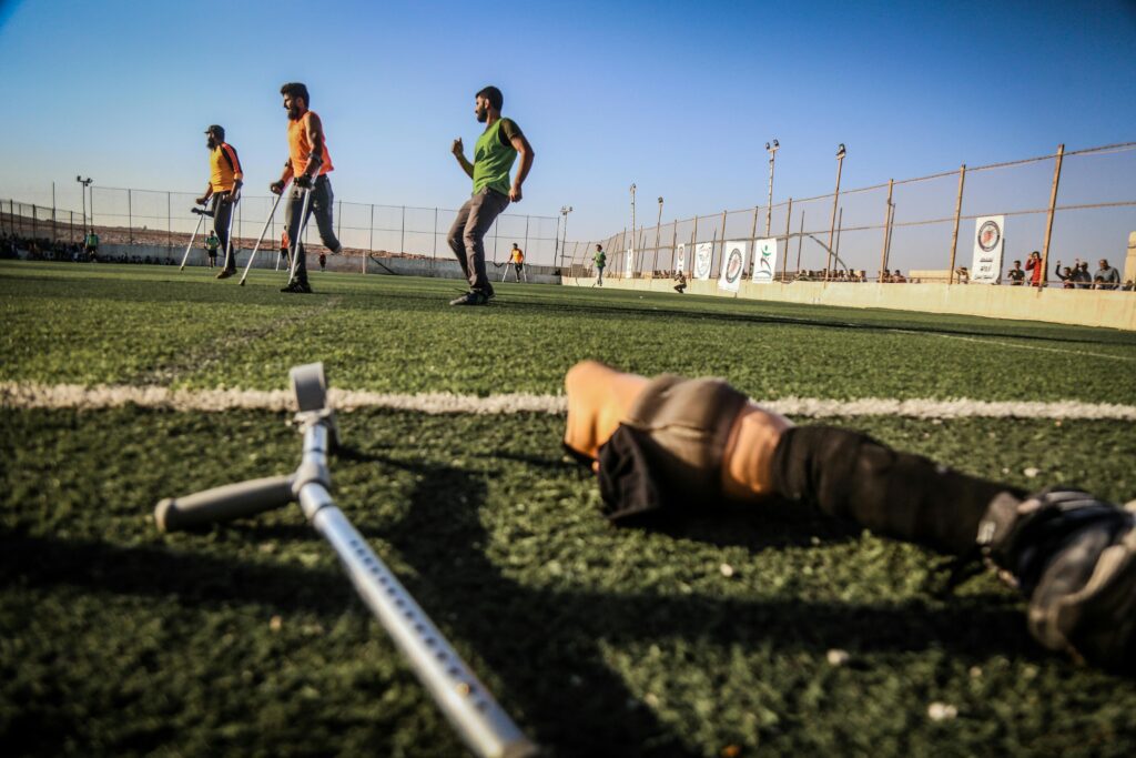 Três homens treinando em uma grama sintética, praticando um esporte com determinação. Cada um possui próteses de metal nas pernas, destacando sua resiliência e força de vontade em superar desafios e se dedicar ao esporte, mesmo diante das dificuldades do processo.