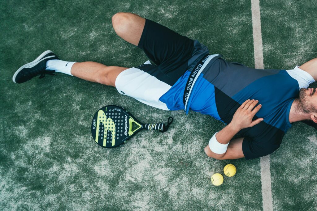 Homem deitado na grama de uma quadra de tênis, aproveitando um momento de descanso. Sua postura reflete resiliência e a compreensão de que pausar para se recuperar é parte essencial do processo de aprendizado e evolução no esporte