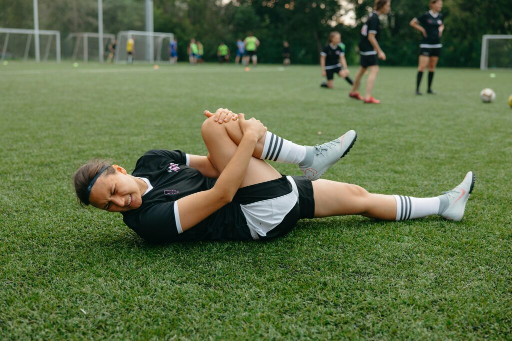 Uma atleta de futebol feminino, caída no chão com a perna direita machucada, expressa dor enquanto luta para superar a lesão. Apesar da dificuldade do momento, ela demonstra a força e determinação necessárias para enfrentar o desafio e continuar sua jornada no esporte.