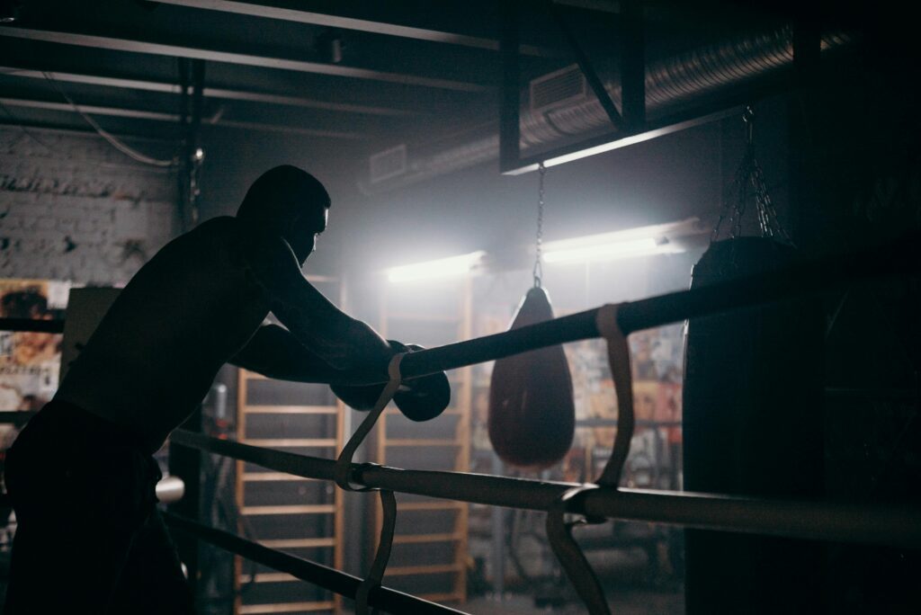 Pugilista na academia, descansando no ringue após um treino intenso, com o braço apoiado nas cordas, em um momento de reflexão e recuperação.
