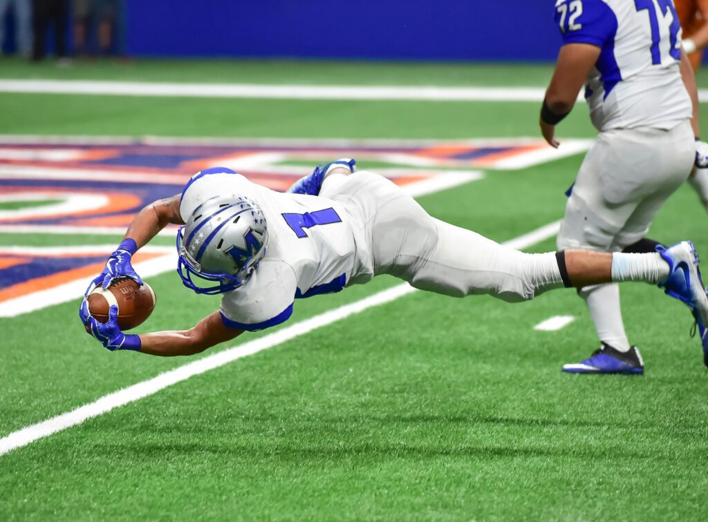Jogador da NFL mergulhando em direção à endzone, segurando firmemente a bola enquanto realiza um peixinho decisivo para marcar o touchdown, simbolizando a conquista de sua meta esportiva.