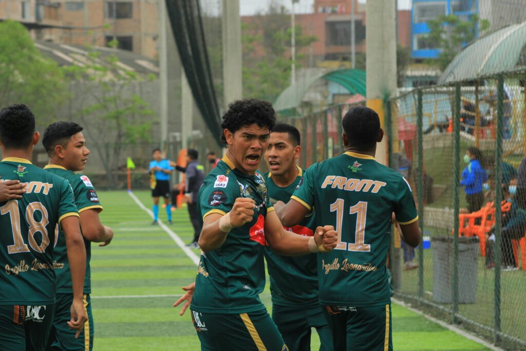 Jogador de futebol em destaque, vibrando intensamente ao comemorar um gol, com expressão de foco e alegria. Ao fundo, seus companheiros se movimentam rapidamente, prontos para reiniciar o jogo e manter a energia da partida.