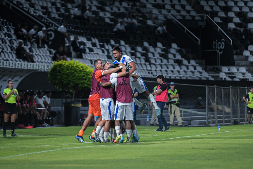 Um jogador titular no centro do campo celebra um gol marcante ao abraçar calorosamente os cinco jogadores reservas, que correram do banco para se juntar à comemoração.