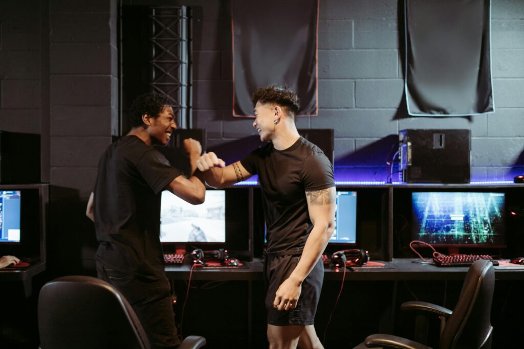 Dois jogadores de eSports, de pé em frente aos computadores, celebram uma vitória com um cumprimento de antebraço, transmitindo felicidade e camaradagem. Suas expressões faciais radiantes refletem o orgulho pela conquista e a conexão construída durante o jogo.