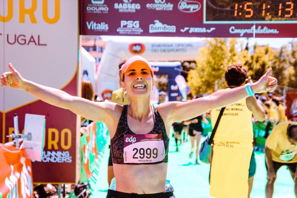 Corredora vibrando de felicidade ao cruzar a linha de chegada, com os braços abertos em celebração e o número de competição estampado no peito, comemorando a conquista de sua meta esportiva de tempo planejada.