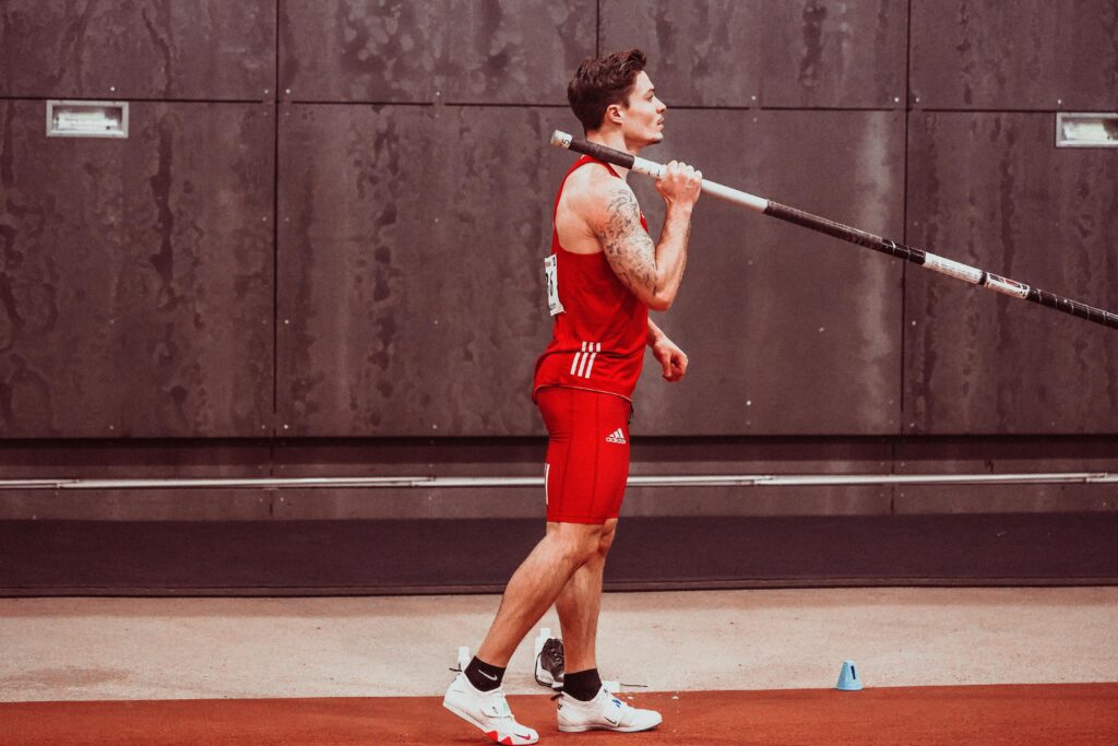 Atleta com a vara em mãos, posicionado na pista de uma arena de competição, concentrado e focado enquanto se prepara para correr e realizar o salto.