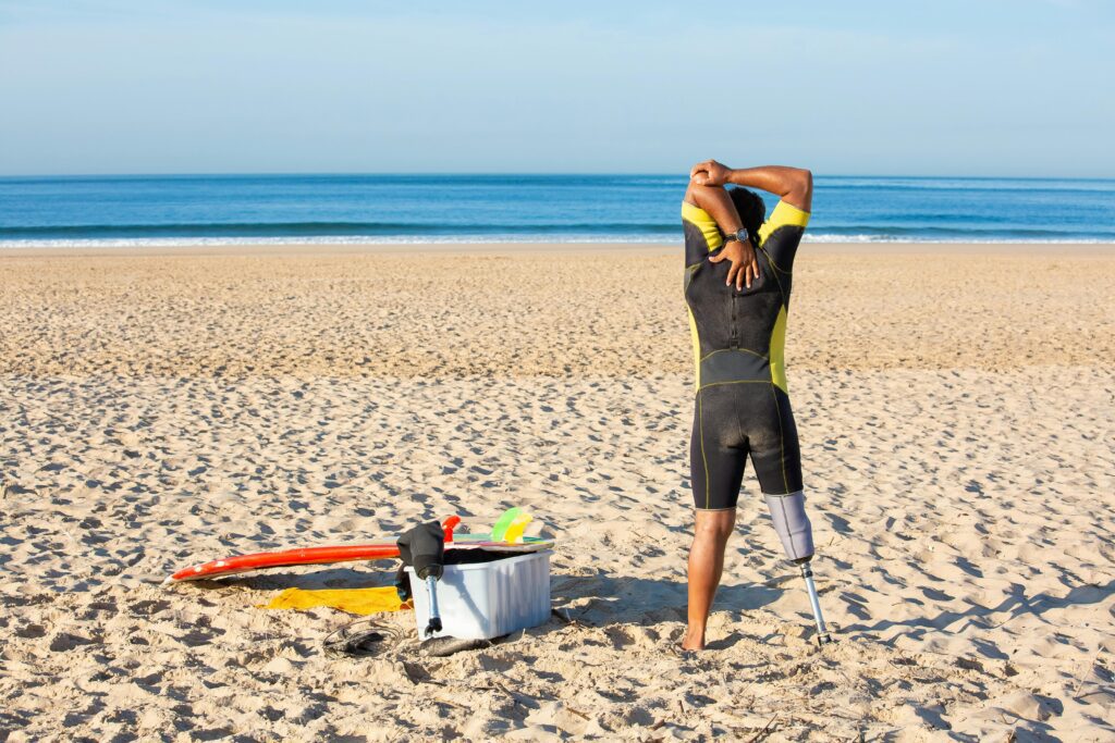 Surfista com uma perna mecânica se alonga à beira do mar, estendendo o braço em preparação para entrar nas ondas, simbolizando superação e resiliência no esporte.
