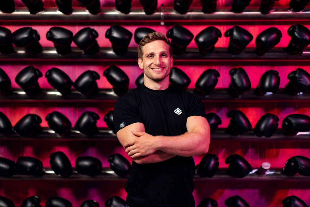 Um Coach de boxe encara a câmera com expressão firme e confiante, cercado por várias luvas ao fundo, transmitindo sua autoestima e segurança na própria experiência e capacidade de treinar grandes atletas.