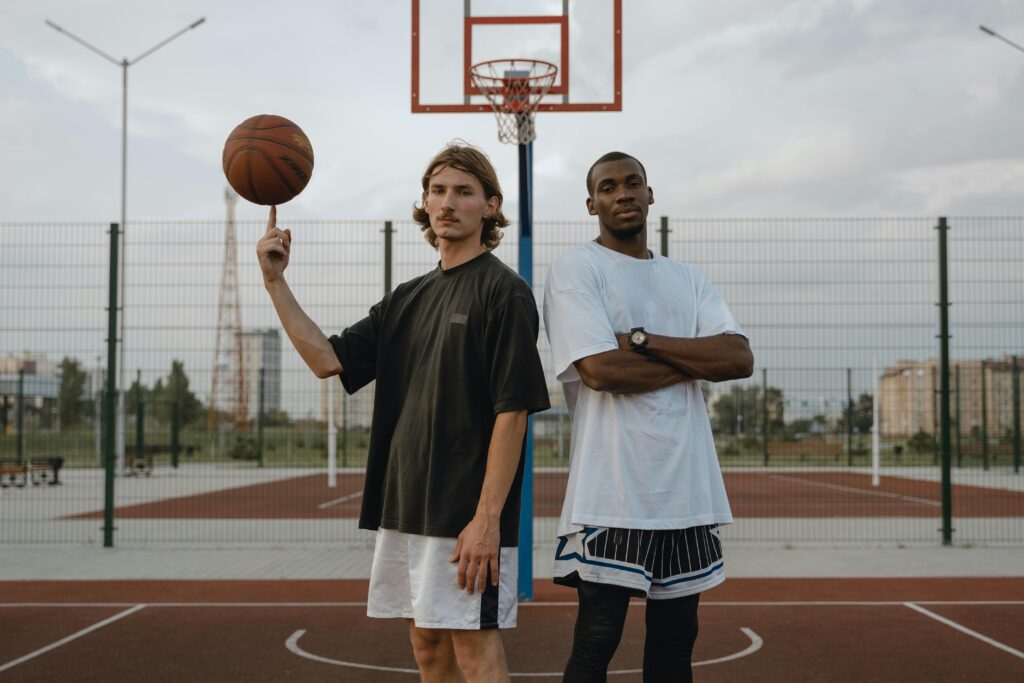Dois atletas de basquete olham para a câmera com foco e seriedade, enquanto um deles equilibra a bola com um dedo, mostrando concentração e habilidade, com o aro ao fundo.
