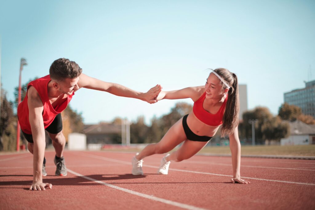 Dois atletas de atletismo, um homem e uma mulher, apoiando-se com um braço no chão enquanto realizam um movimento de prancha, mantendo as pernas esticadas e equilibrando-se apenas com o suporte do outro atleta.