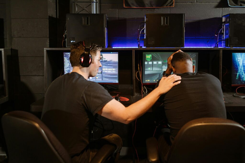 Dois jogadores de esports sentados em frente ao computador; um deles aparenta estar visivelmente chateado, enquanto o outro o consola, colocando o braço sobre seu ombro em um gesto de apoio.