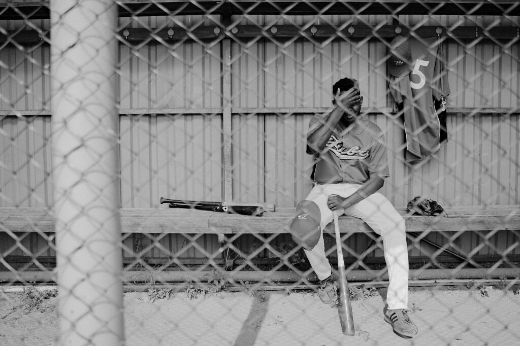 Jogador de beisebol sentado sozinho no banco, cabisbaixo e visivelmente frustrado após perder um lance crucial, capturado em uma imagem em preto e branco com tonalidade melancólica.