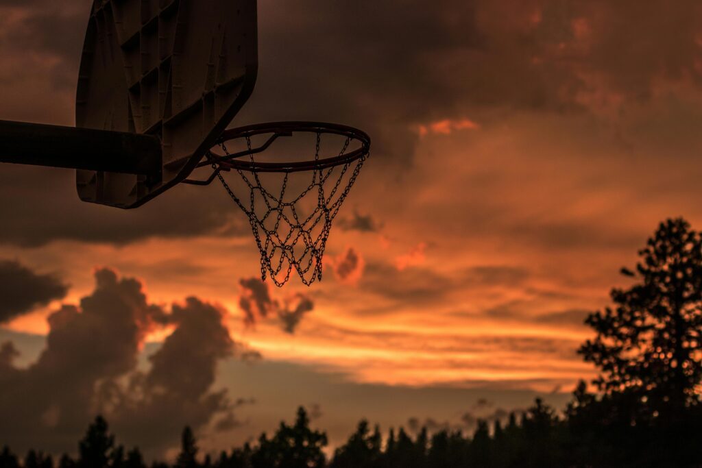 Um aro de basquete em destaque ao entardecer, com o céu tingido em tons alaranjados, criando uma atmosfera tranquila e inspiradora, ideal para aliviar o estresse dos atletas e proporcionar um momento de reflexão no esporte.