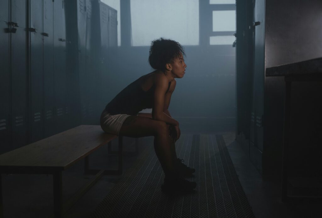 Mulher sentada em um banco no vestiário esportivo, refletindo sobre sua trajetória no esporte e seu progresso até o momento.