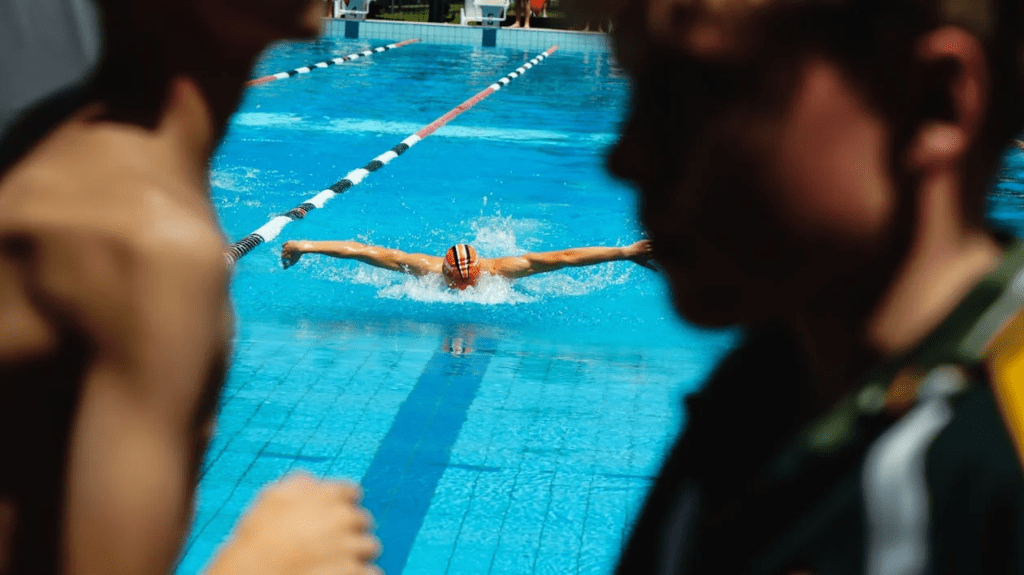Nadador executando a técnica borboleta em uma piscina olímpica, demonstrando força e precisão durante um treino intenso. Na frente da imagem, duas pessoas passam, criando uma sensação de movimento e profundidade na cena.