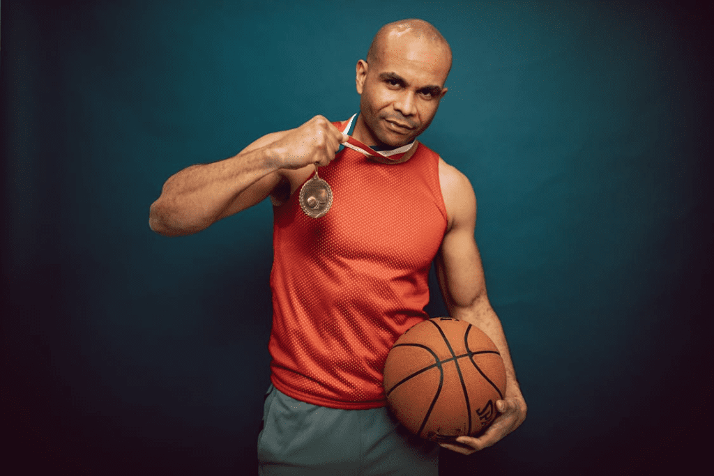 Um jogador de basquete com uma medalha de ouro no pescoço, segurando-a com uma mão enquanto mantém a outra firme em uma bola de basquete. Seu olhar é determinado, transmitindo foco e a mentalidade vencedora que o levou à conquista.