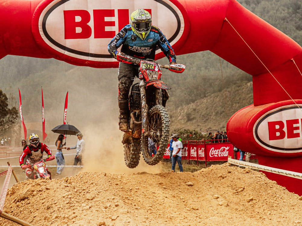 Um atleta de motocross salta sobre um declive, cruzando a linha de chegada com concentração total. Seu traje coberto de lama reflete os desafios da prova, simbolizando aprendizado e resiliência após quedas anteriores.