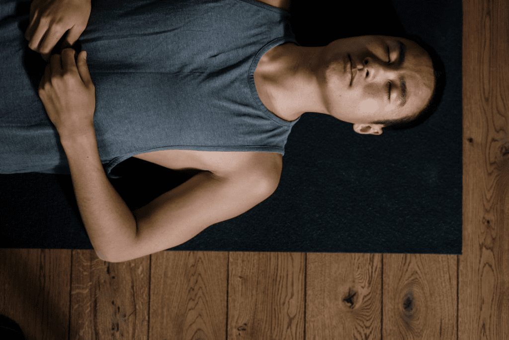 Um homem está deitado em um colchonete, exausto após uma sequência intensa de treino. Com a respiração ofegante e o olhar fixo no teto, ele aproveita o momento para recuperar as energias, sentindo o peso do esforço e a satisfação do dever cumprido.