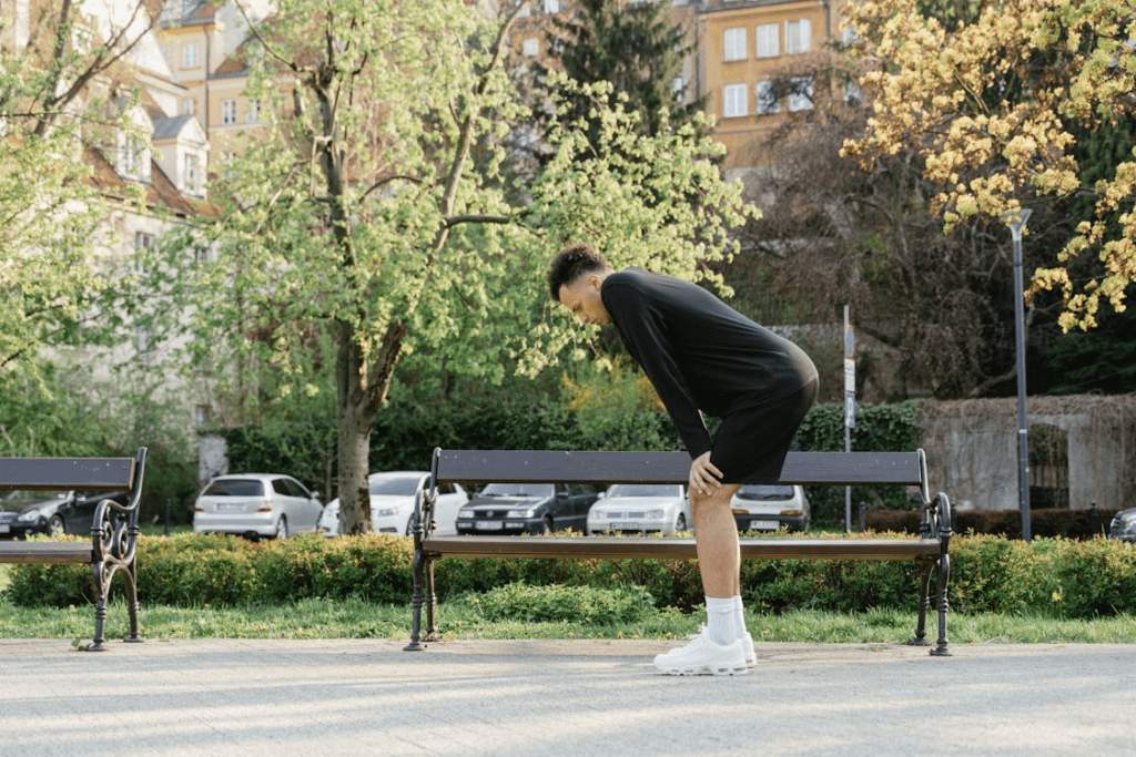 Um homem, com as mãos apoiadas nas pernas e a coluna inclinada para frente, descansa após uma corrida no parque. Com um banco ao fundo e vestindo roupa esportiva, ele recupera o fôlego enquanto reflete sobre seu desempenho.
