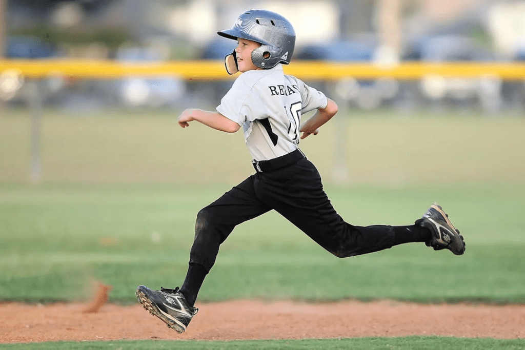 Criança atleta de beisebol em plena corrida entre bases, demonstrando percepção e tomada de decisão ao identificar o momento certo para avançar e completar o circuito. Um gesto de coragem, estratégia e aprendizado em ação.