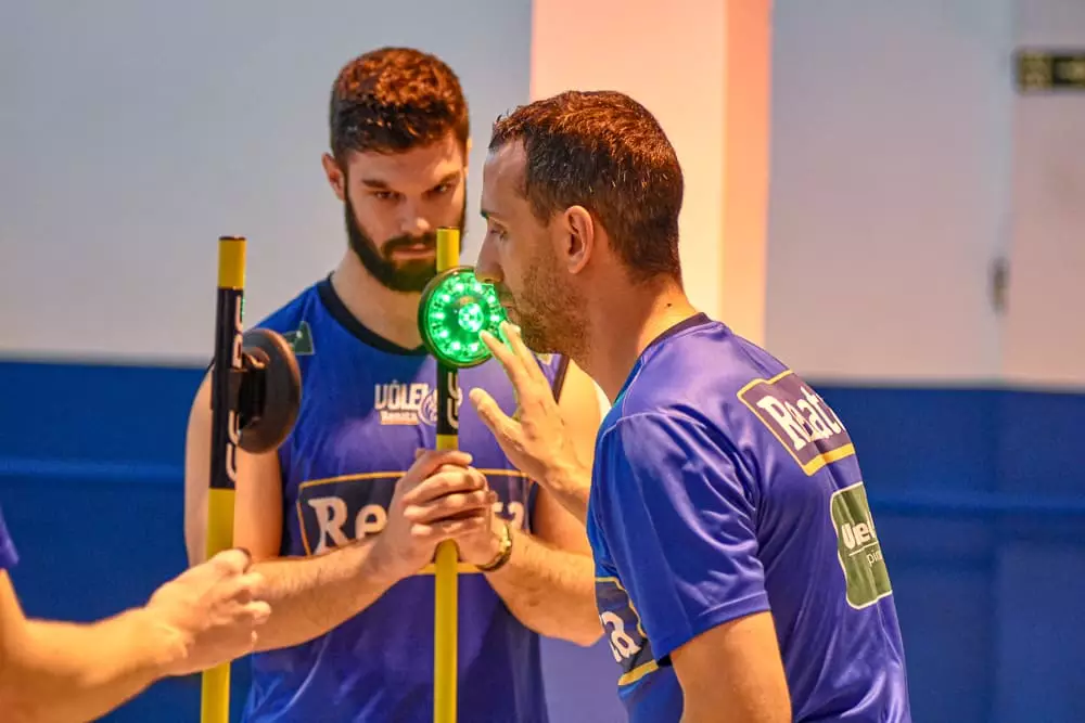 Atleta de vôlei realizando um treino cognitivo com foco em agilidade e tempo de reação, tocando luzes que piscam rapidamente enquanto outro atleta as segura. A atividade estimula concentração, velocidade de resposta e tomada de decisão em situações de alta pressão.