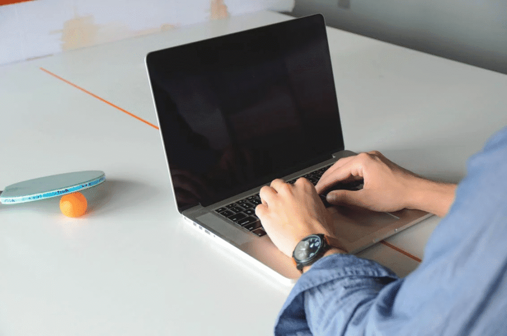 Pessoa de costas, trabalhando em um notebook sobre uma mesa de ping pong, com uma raquete apoiada e segurando uma bolinha, simbolizando a conexão entre os mundos do esporte e do ambiente corporativo.