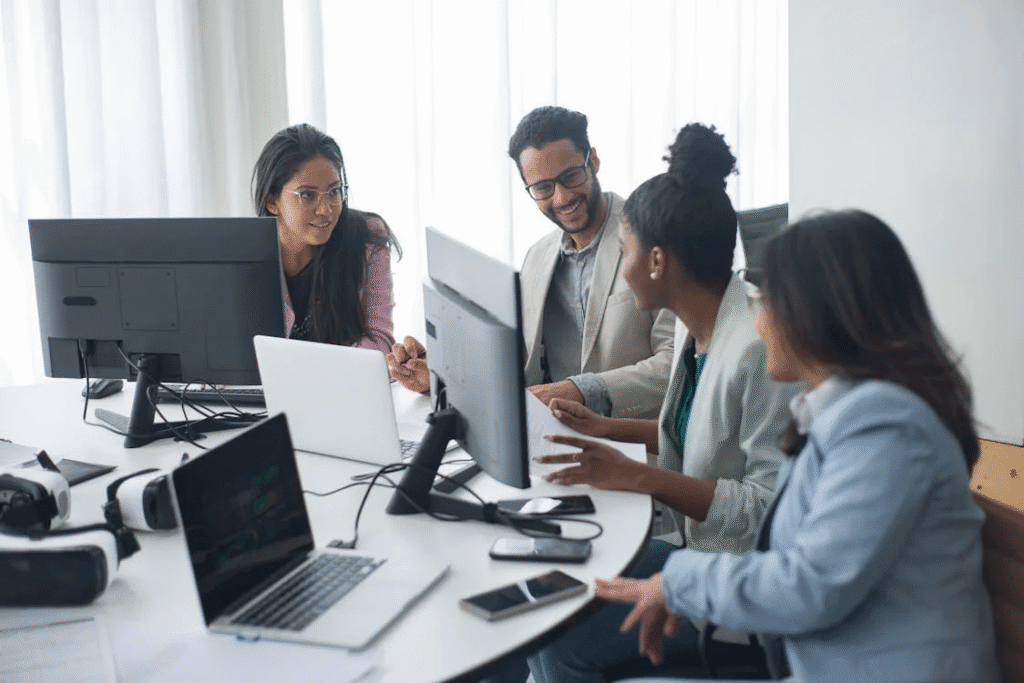 Quatro pessoas vestidas de social, lado a lado em um escritório, com computadores à frente, colaborando e se apoiando em um dia de trabalho organizacional.