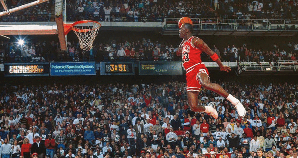 Michael Jordan salta em direção ao aro, prestes a enterrar, enquanto a torcida assiste atenta em um estádio lotado.