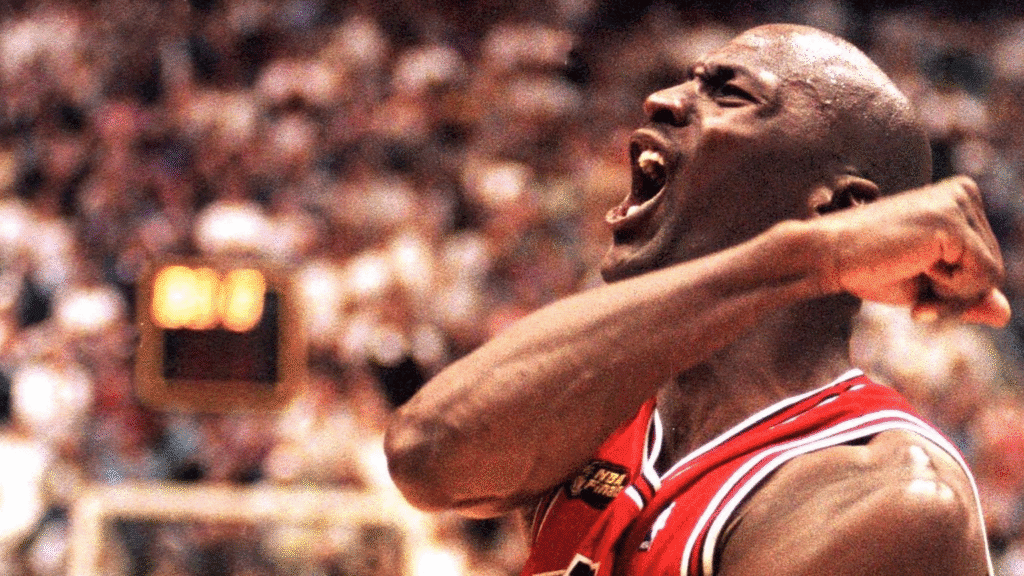 Michael Jordan celebra com o punho cerrado e grito de vitória, vibrando junto à torcida após marcar um ponto decisivo.