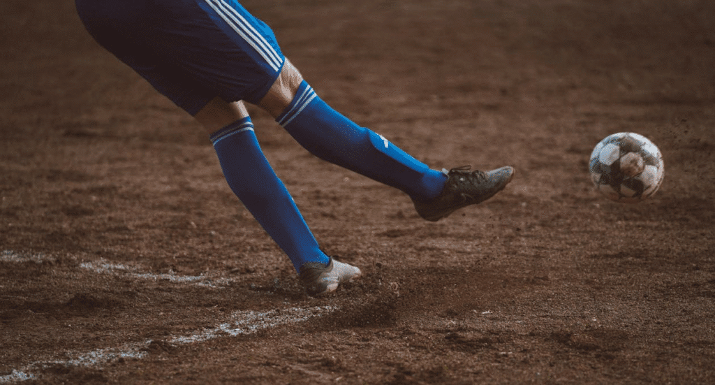 Jogador de futebol em campo de terrão chuta a bola com força após cobrança de falta, demonstrando intensidade e determinação no jogo.