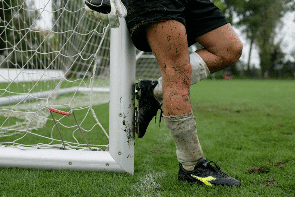 Goleiro em campo, capturado da cintura para baixo, com chuteira coberta de grama, demonstrando intensidade na entrega da sua transição ao profissional enquanto prepara a barreira e apoia um dos pés na trave durante uma cobrança de falta.