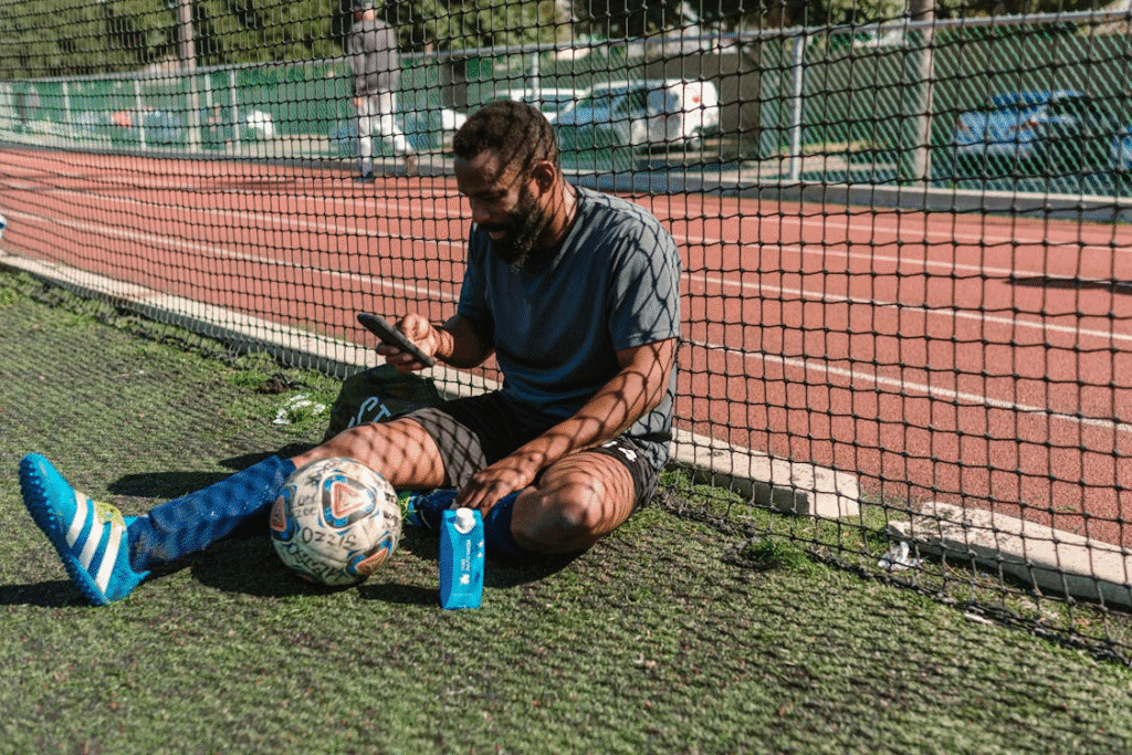 Homem trajado com uniforme de futebol, sentado na grama e olhando para o celular, analisando comentários e notícias sobre sua performance nas redes sociais.