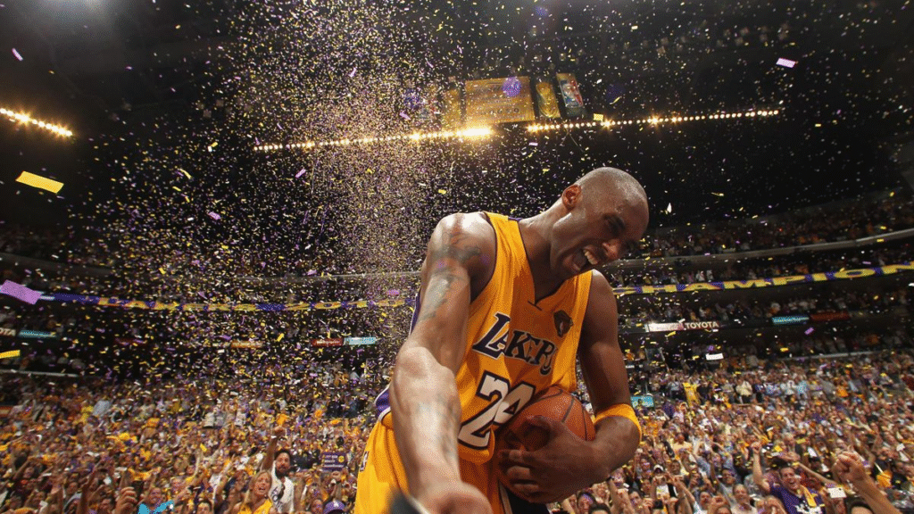 Kobe Bryant comemorando a conquista de um título da NBA, cercado por papel picado e com a torcida do Los Angeles Lakers ao fundo, dentro do estádio. A imagem simboliza a consagração da dedicação e mentalidade vencedora de um dos maiores atletas da história do basquete.