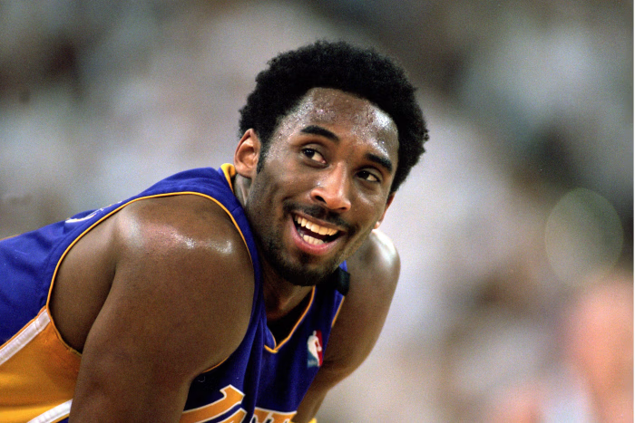 Kobe Bryant, ainda com cabelo, apoiado nas coxas e sorrindo para o público durante uma partida da NBA. Mesmo visivelmente cansado, sua expressão transmite alegria e paixão pelo basquete, refletindo sua resiliência e conexão com os fãs.