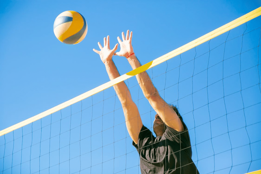 Jogador de vôlei de praia executando um bloqueio preciso, saltando no momento exato para interceptar a bola que vem em sua direção. A imagem destaca a tomada de decisão rápida, o tempo de reação e a técnica defensiva essencial no alto nível do esporte.