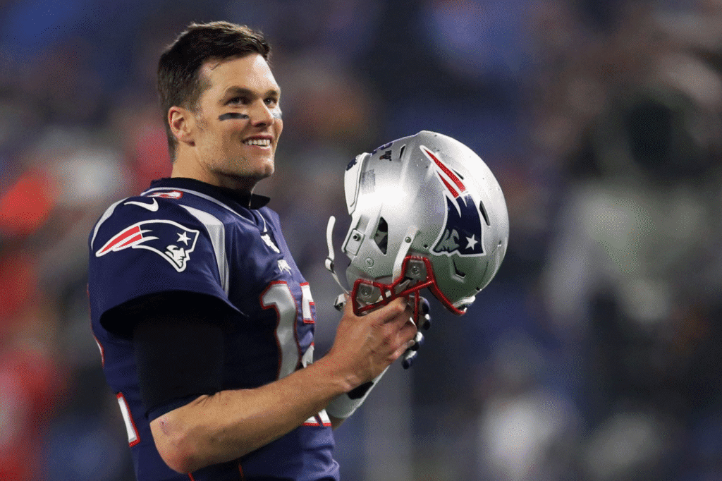 Tom Brady durante um jogo pelos Patriots, segurando o capacete nas mãos e sorrindo ao olhar para o alto, logo após realizar uma jogada de destaque na partida.