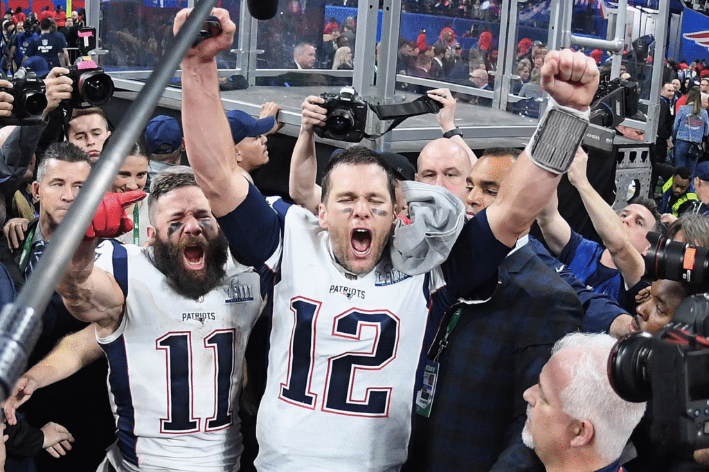Tom Brady celebra a conquista de um título pelo New England Patriots, com o braço erguido e olhos fechados, rodeado por colegas de equipe, imprensa e torcedores em êxtase.
