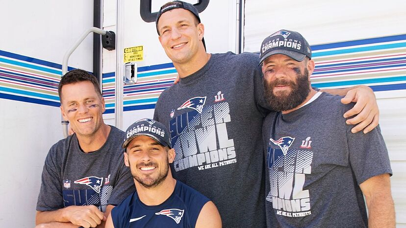 Tom Brady ao lado de amigos do esporte, vestindo uma camisa comemorativa dos títulos conquistados pelos Patriots, em um momento descontraído e de celebração da carreira.