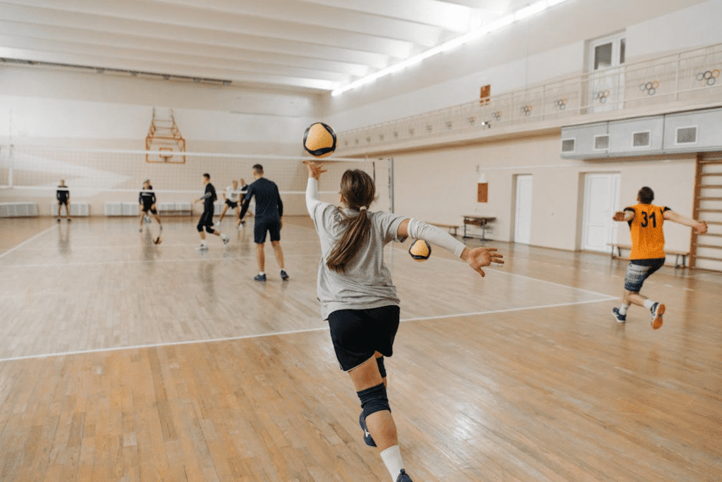 Treino de vôlei em quadra, com atleta se preparando para sacar enquanto diversas práticas e exercícios acontecem ao redor.