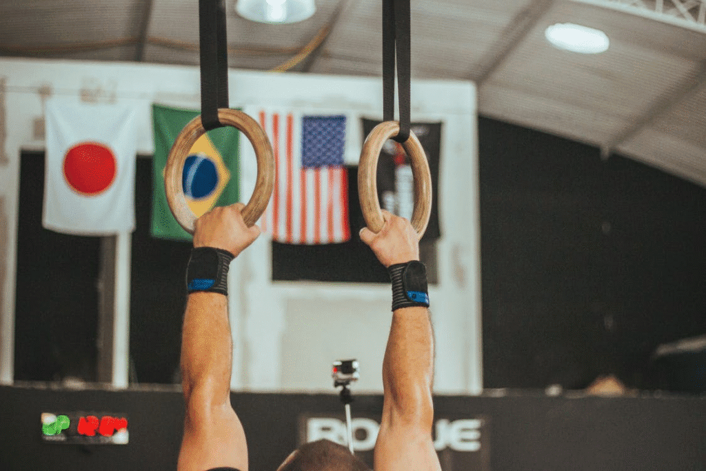 Imagem de um ginasta segurando firmemente as argolas, com foco apenas nas mãos. Ao fundo, bandeiras de diversos países indicam o cenário de uma competição de nível internacional.