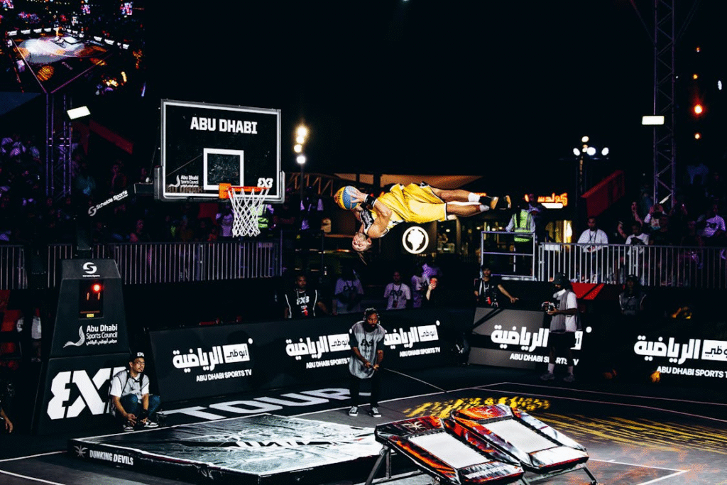 Atleta de basquete acrobático em ação, utilizando um trampolim para realizar manobras aéreas antes de enterrar a bola, demonstrando habilidade e explosão física em altura elevada.