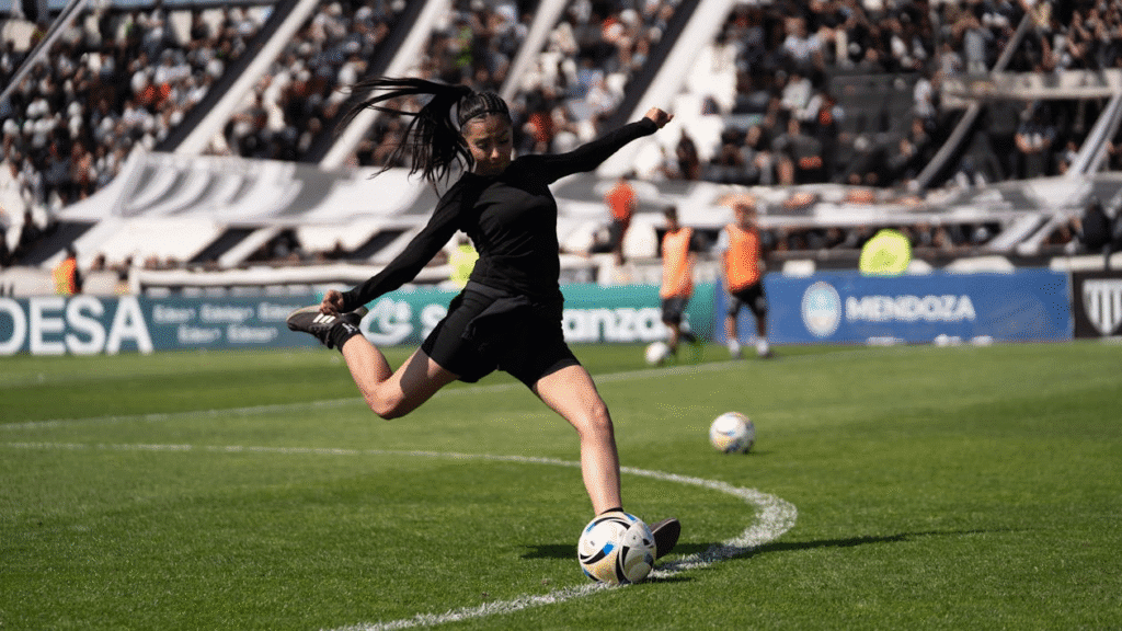 Jogadora de futebol com camiseta preta realiza um chute durante o aquecimento pré-jogo, dentro de um estádio com a torcida ao fundo, demonstrando preparação e foco antes da partida.