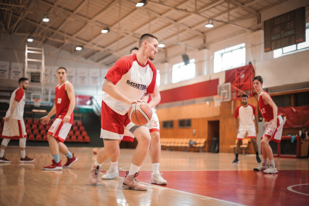 Equipe de basquete dentro da quadra iniciando os treinos com alegria e entrosamento. O grupo pratica drills e jogadas coletivas, mostrando cooperação e espírito esportivo durante a atividade.