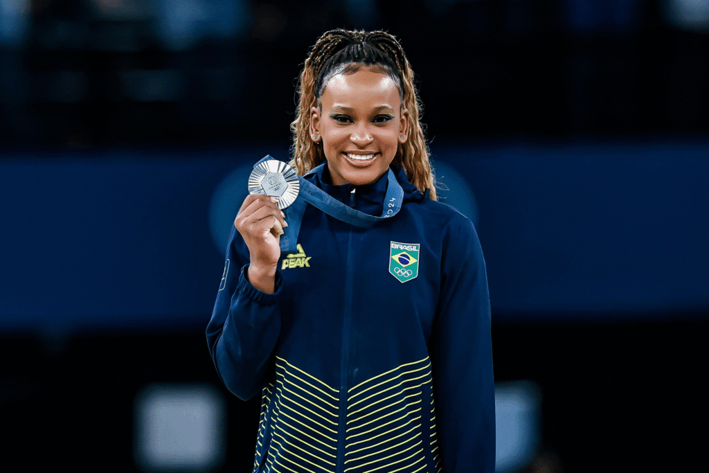 Rebeca Andrade no pódio segurando a medalha de prata conquistada por equipe nos Jogos Olímpicos de Paris 2024, sorrindo com orgulho ao lado das colegas do Time Brasil.