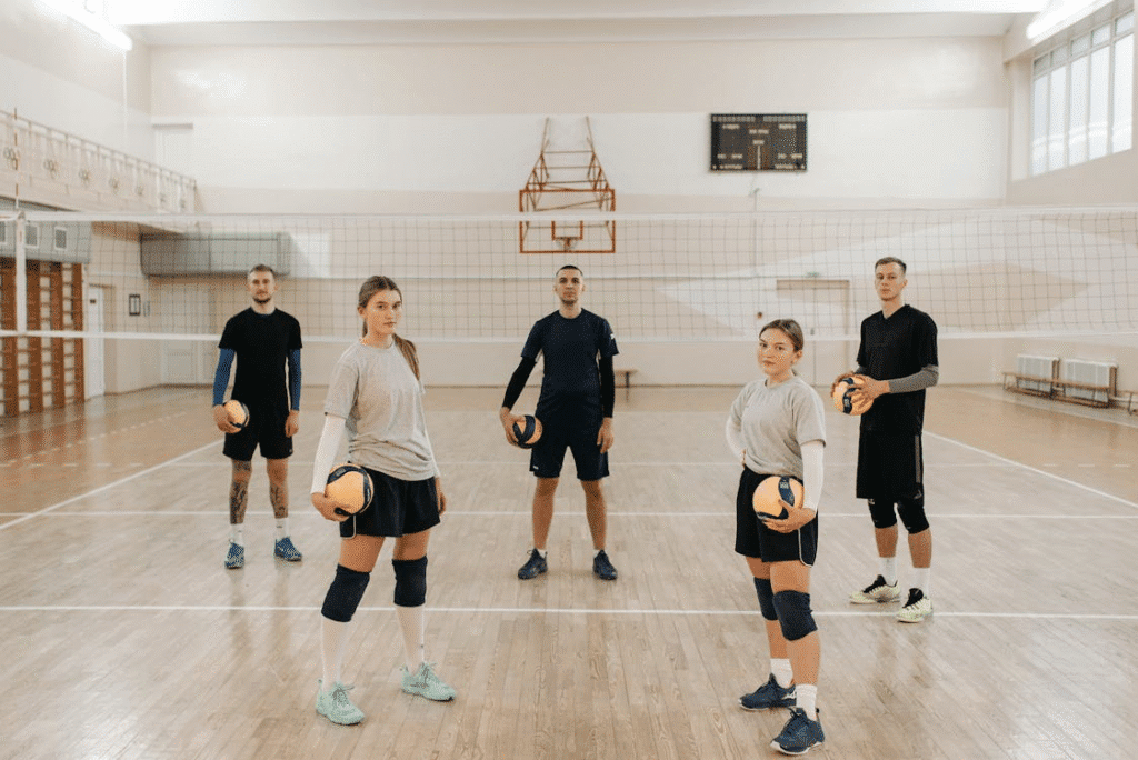 Time de vôlei posando para foto durante o treino, todos do mesmo lado da rede e segurando bolas de voleibol, em uma quadra vazia. O grupo sorri para a câmera, transmitindo união e dedicação ao esporte.