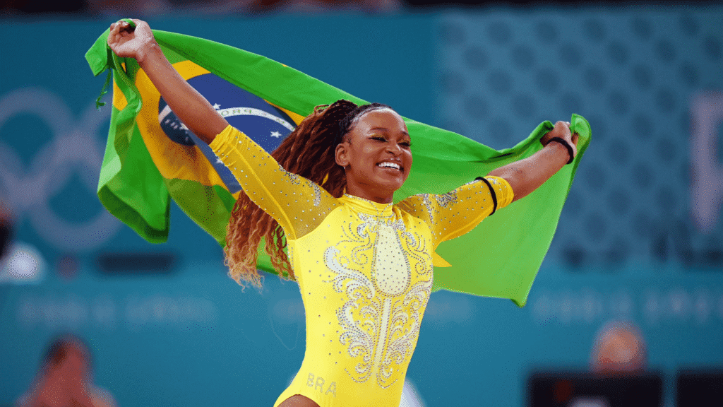 Rebeca Andrade radiante, vestida com o uniforme verde, amarelo e branco do Brasil, segurando a bandeira do país com os braços abertos após mais uma conquista internacional.