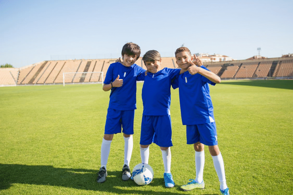Três meninos por volta de 12 anos treinando futebol em um campo gramado, sorridentes e abraçados, com a bola nos pés e o céu aberto ao fundo, simbolizando amizade e diversão no esporte.