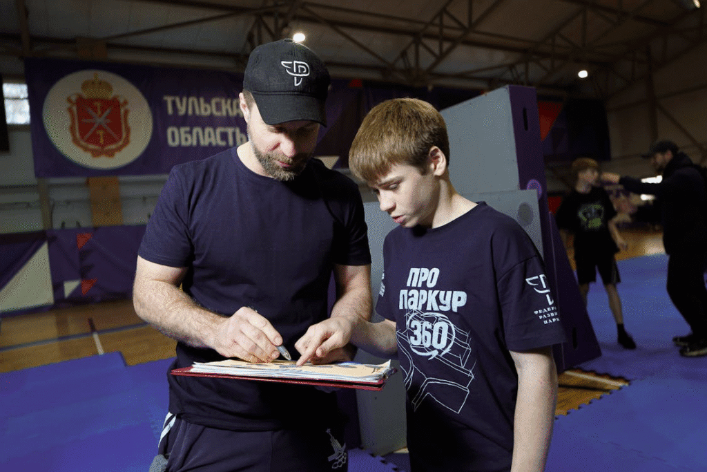 Em um tatame, um atleta de luta mostra em um manual técnico como executar uma movimentação correta. O outro atleta observa atentamente, buscando entender a tática para melhorar a performance. A cena representa disciplina e estudo além do físico.