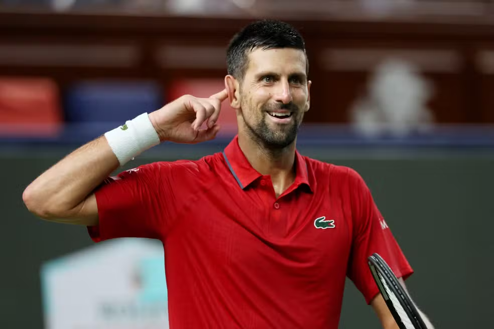 Foto de Novak Djokovic sorrindo e incentivando a torcida, colocando o dedo no ouvido para ouvir seu nome após um ponto importante. Cena clássica do sérvio, conhecida pela intensidade emocional e mentalidade competitiva. Excelente para artigos sobre força mental, controle emocional e cultura do tênis.