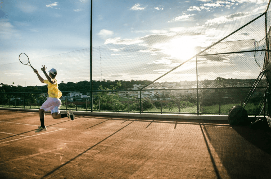 Imagem de um jogador de tênis amador tentando retornar a bola em pleno salto, sob sol forte e clima quente. O cenário tem quadra aberta, bastante vegetação ao redor e iluminação intensa, representando esforço físico, lazer esportivo e prática do tênis como atividade de saúde e bem-estar. Ideal para textos sobre tênis amador, fundamentos técnicos e condicionamento físico.